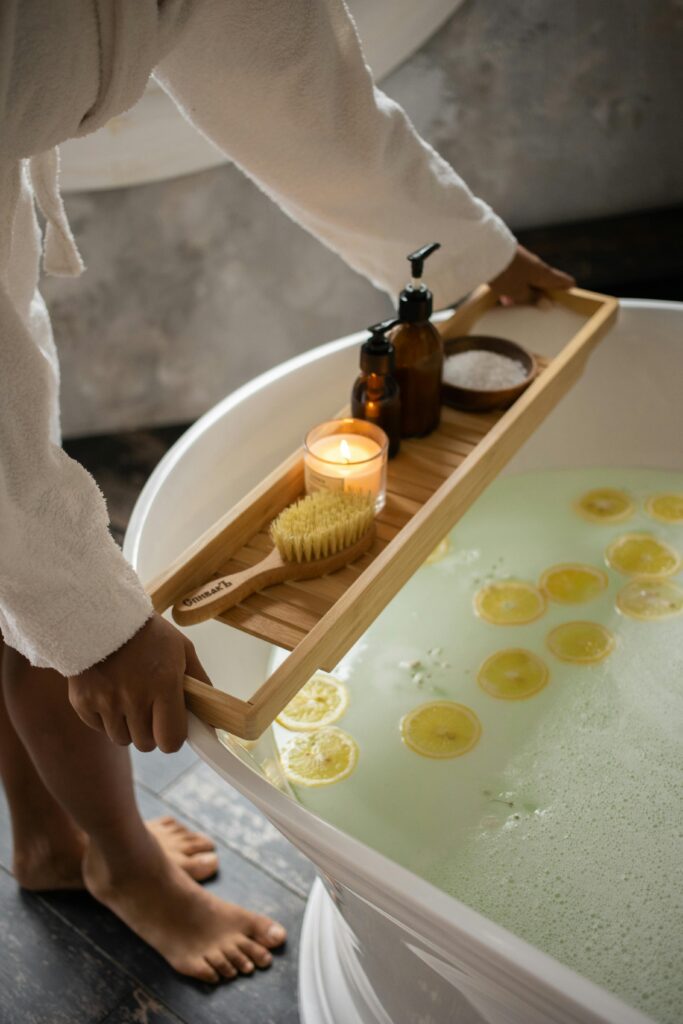 female in bathrobe putting wooden tray with spa supplies on tub with lemon slices on water surface during skincare routine