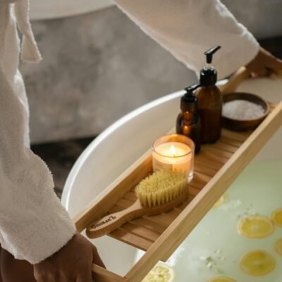 female in bathrobe putting wooden tray with spa supplies on tub with lemon slices on water surface during skincare routine