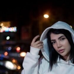 A young woman in a white hoodie poses outdoors at night.