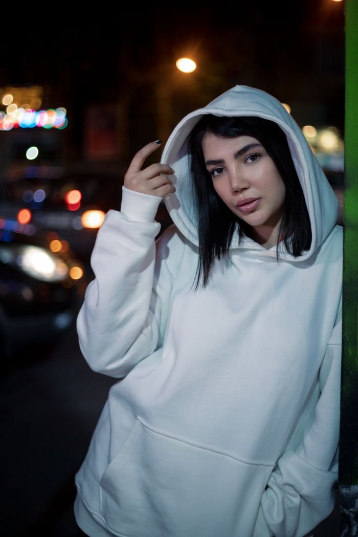 A young woman in a white hoodie poses outdoors at night.